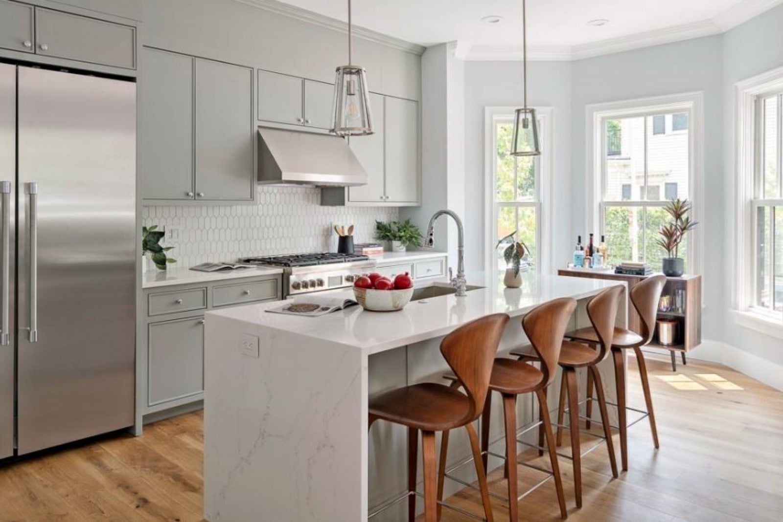 A kitchen with smooth-front gray cabinetry that goes to the ceiling, an island with waterfall edges and seating for four, hardwood flooring, two pendant lights, and a windowed breakfast nook. This photo is used to illustrate open houses going on this weekend.